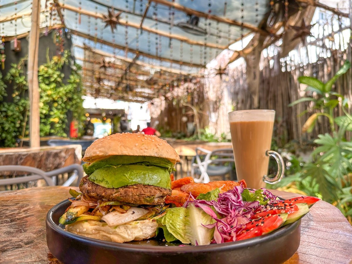 A vibrant vegan burger topped with avocado, served with a side of salad and sweet potato fries, alongside a latte, set in the lush, plant-filled outdoor seating area of Arte Sano on Holbox Island.