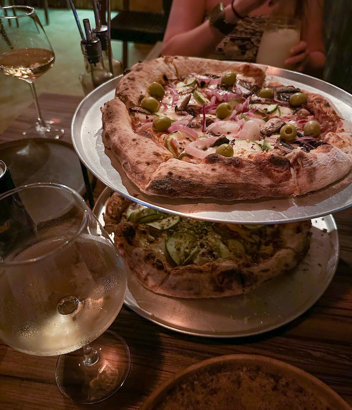 Two freshly baked pizzas topped with olives, onions, and vegetables, served on metal trays with a glass of white wine at Crónicas Taproom on Holbox Island.