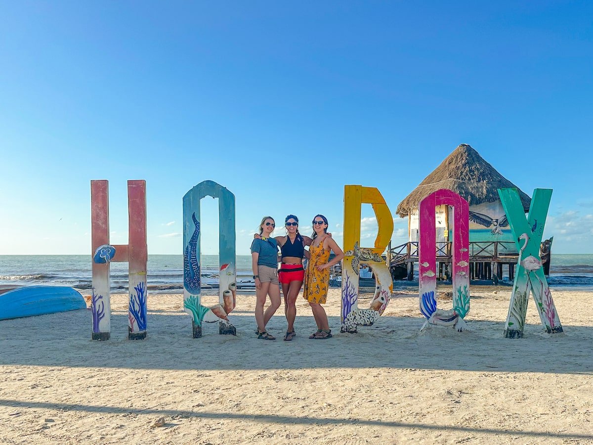 Three women posing in front of the colorful 'Holbox' sign on the beach, with a thatched hut and the ocean in the background under a bright blue sky.