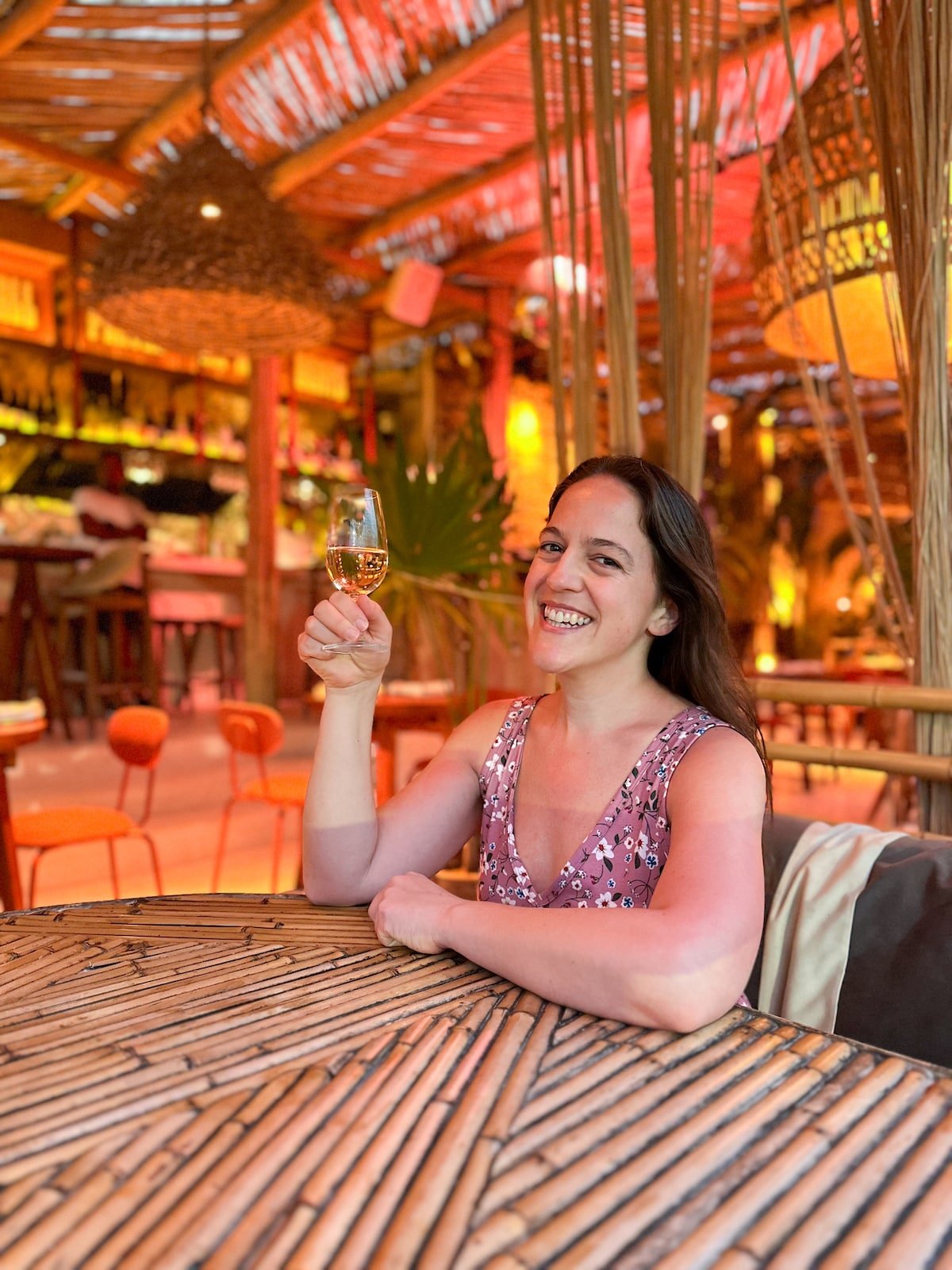 Smiling woman in a floral dress holding a glass of wine at Lazaro Wine Bar on Holbox Island, surrounded by warm lighting and cozy, rustic decor.