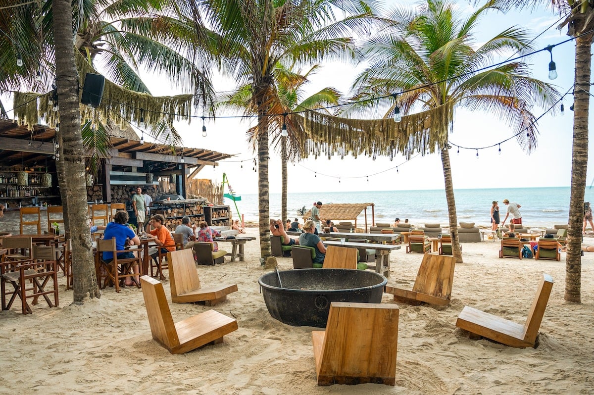 Dining area of Mandarina Restaurant on Playa Holbox, featuring wooden chairs and tables set directly on the sand, surrounded by palm trees and string lights, with a view of the ocean in the background.