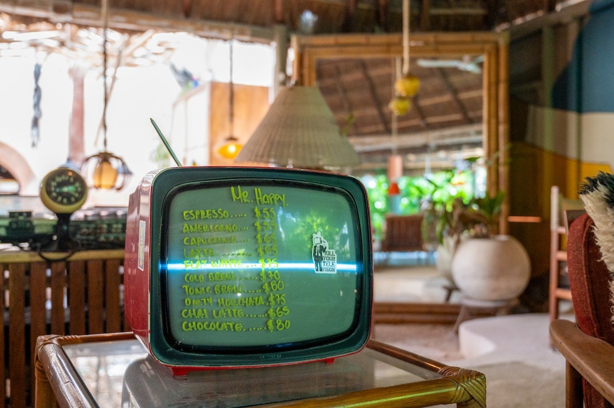 Vintage TV displaying the coffee menu at Mr. Happy Holbox Cafe on Holbox Island, listing espresso, cappuccino, latte, and other drink options, with a cozy, retro-themed cafe setting in the background.