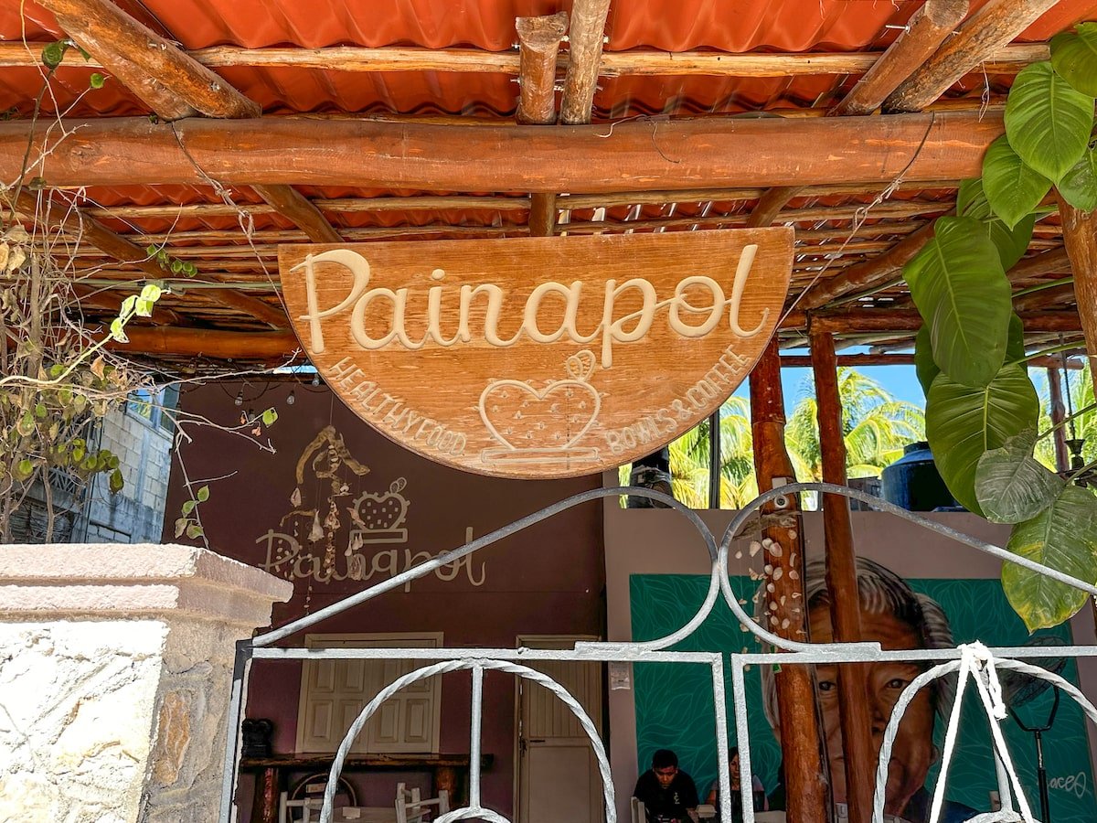 Wooden sign for Painapol cafe in Holbox, reading 'Painapol - Healthy Food, Bowls & Coffee,' hanging under a rustic, wood-beamed roof with lush green plants in the background.