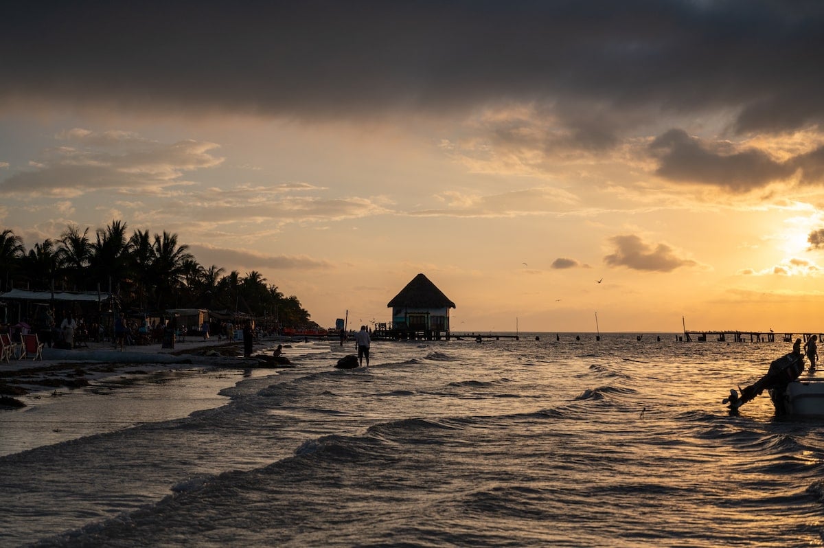 Sunset on Playa Holbox with soft waves reaching the shore, silhouettes of people along the beach, and a thatched hut on stilts over the water under a dramatic sky.