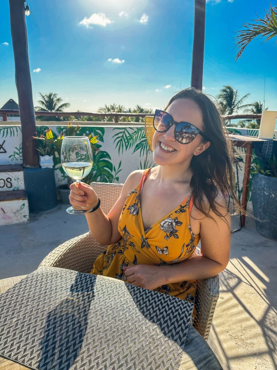 Smiling woman in a yellow floral dress holding a glass of white wine at Soulbox by Mittoz, a rooftop bar on Holbox Island, with tropical plants and palm trees in the background.